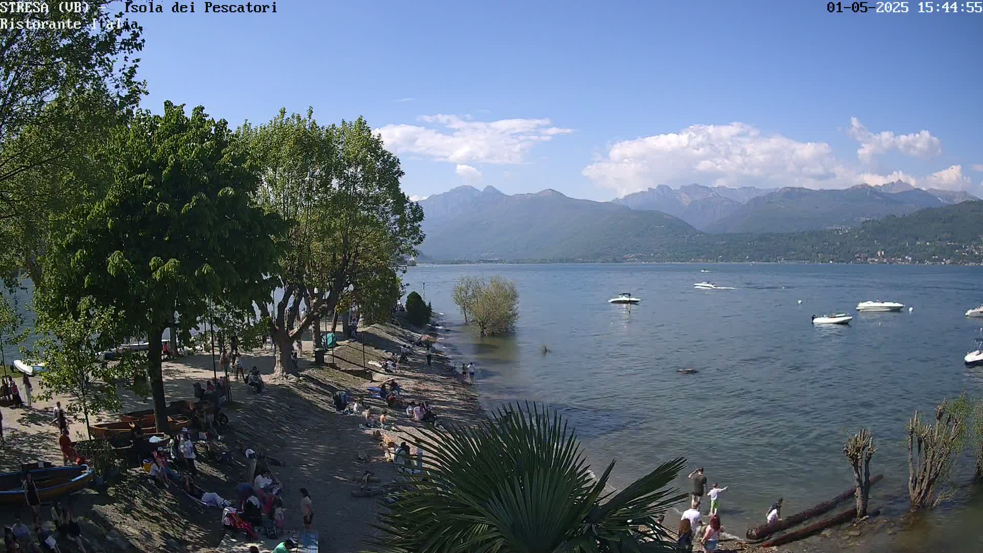 A sunny day at a lakeside beach, showing people relaxing and boats on calm water with mountains in the background.