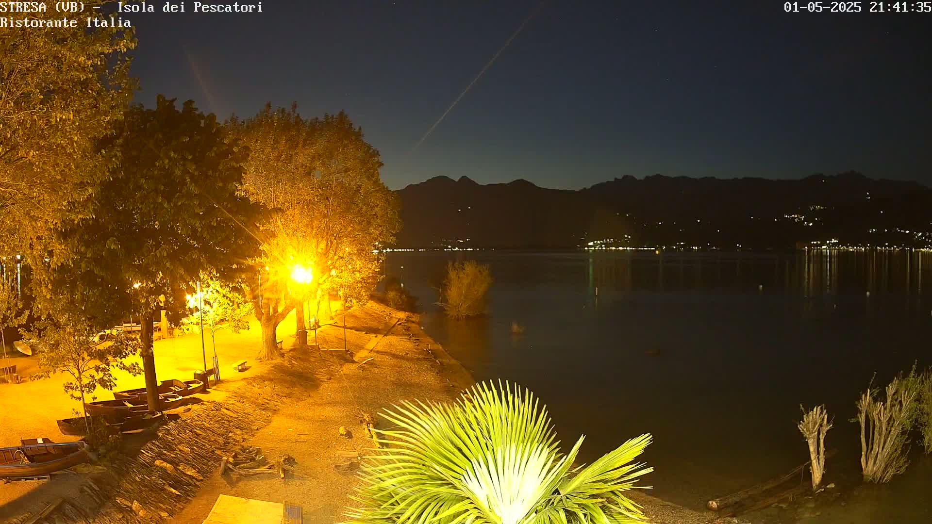 A nighttime view of a calm lake under a clear, dark sky, with illuminated trees and boats on the shore, and mountains in the distance.