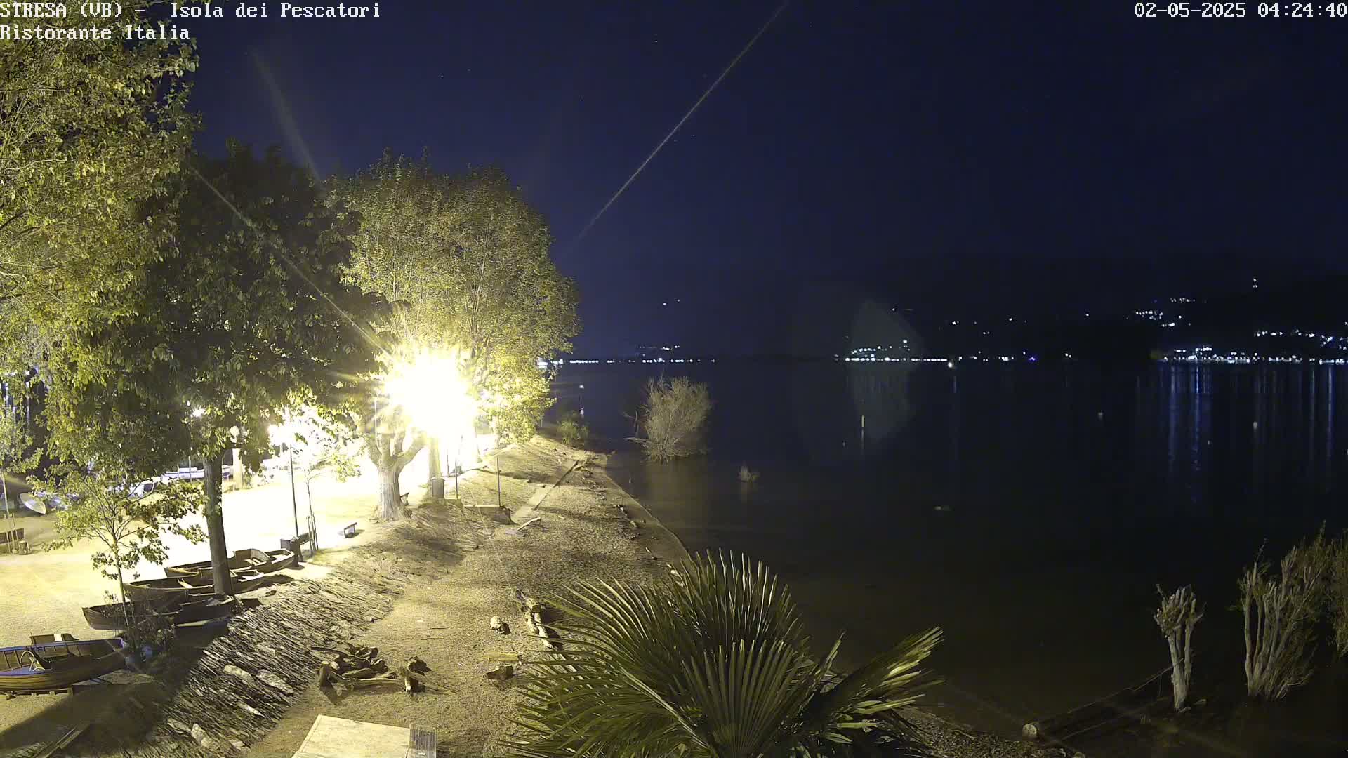 At night, a shoreline with trees, lights, and several small boats is visible beside a calm, dark lake reflecting distant lights.
