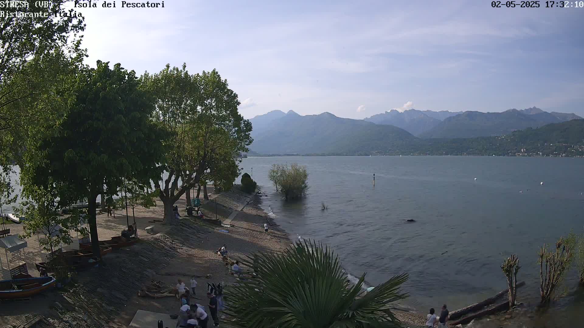 A calm lake with mountains in the background, under a partly sunny sky, is viewed from a lakeside area with several people near some trees and boats.