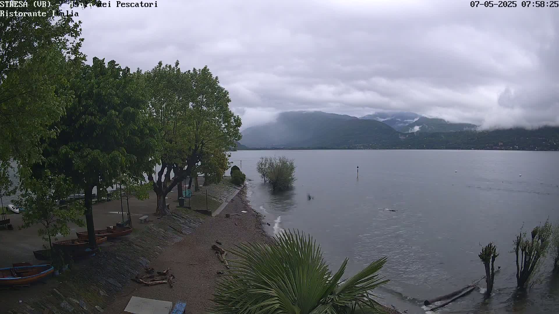 A lakeshore scene under a cloudy sky shows several boats pulled ashore near leafy trees, with calm water lapping the pebbled beach and mountains in the distance.