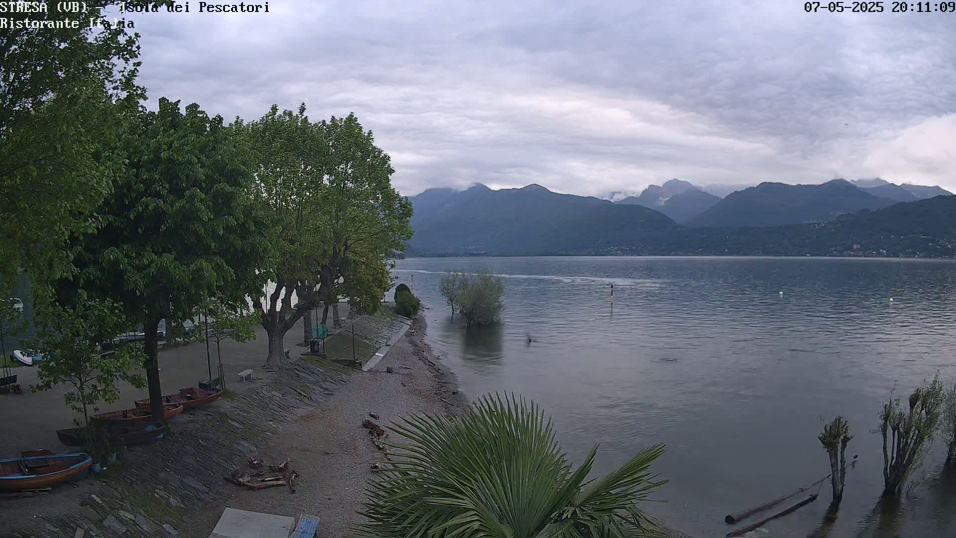 Overcast skies hang over a calm lake, nestled beside a shoreline with several small boats and lush green trees, mountains rising in the distance.