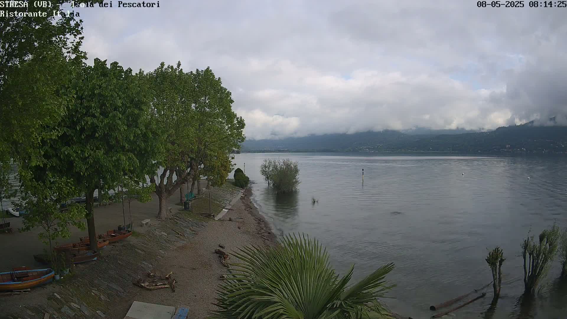 A lakeside scene on a cloudy day shows a pebble beach with several small boats and trees, with calm water extending to distant, fog-covered mountains.