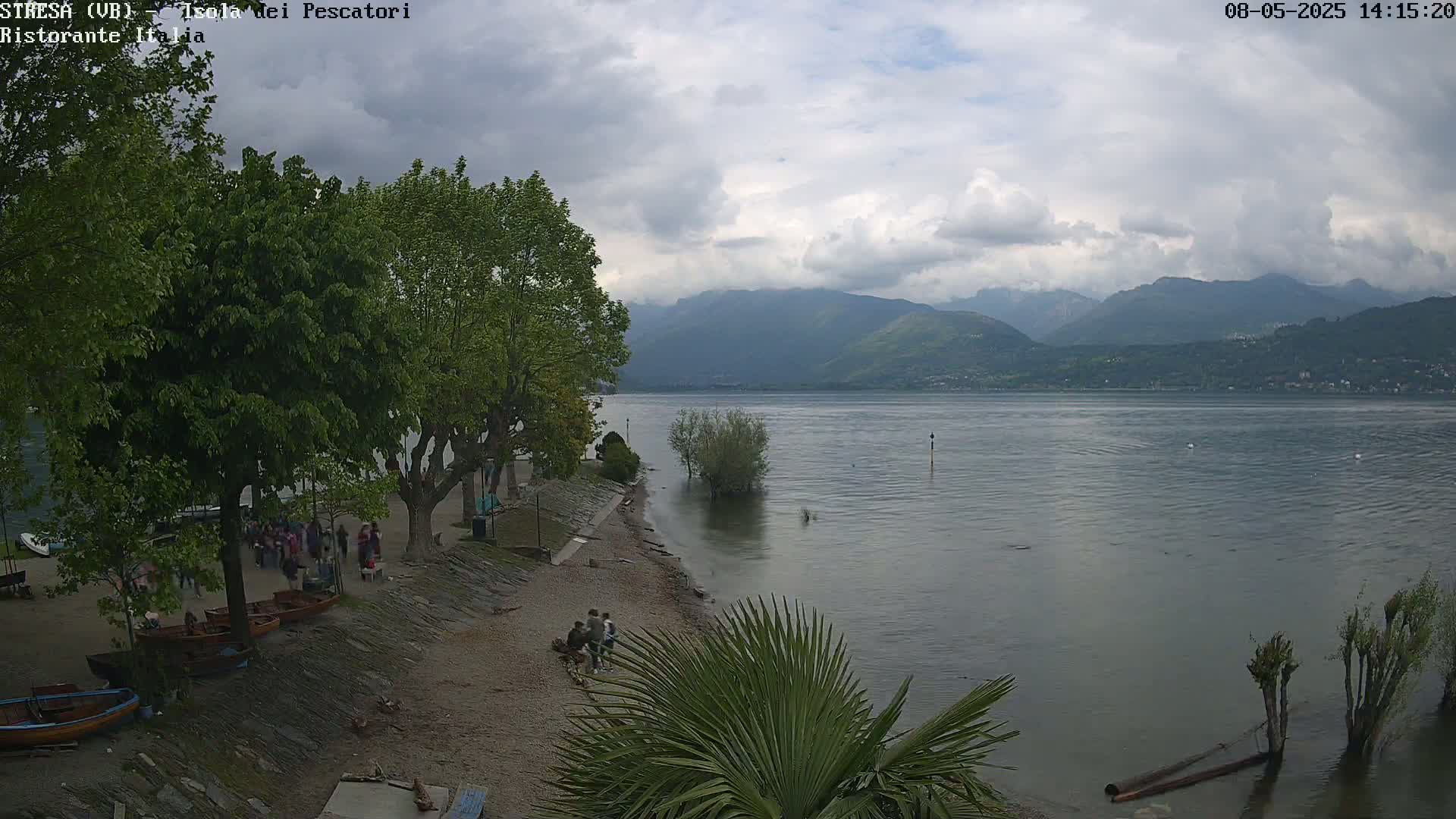 A partly cloudy day shows a calm lake with mountains in the background, a sandy shore with several people and small boats, and leafy green trees lining the shoreline.