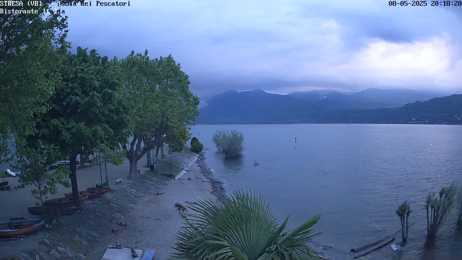 A pebble beach with several small boats and trees overlooks a calm lake under a cloudy sky, with mountains in the distance.