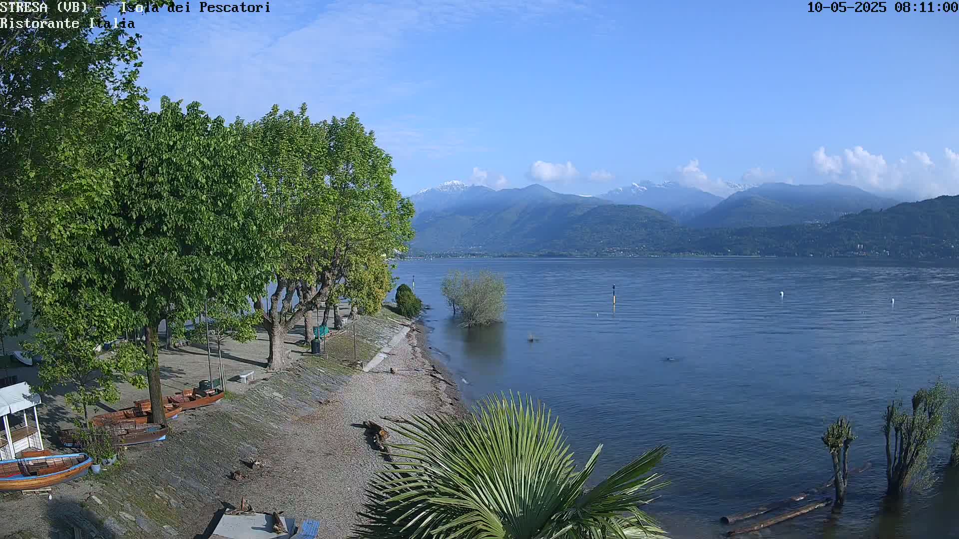 A calm lake with a pebbled shore lined with trees and small boats, under a mostly clear sky, backed by distant mountains with some snow.