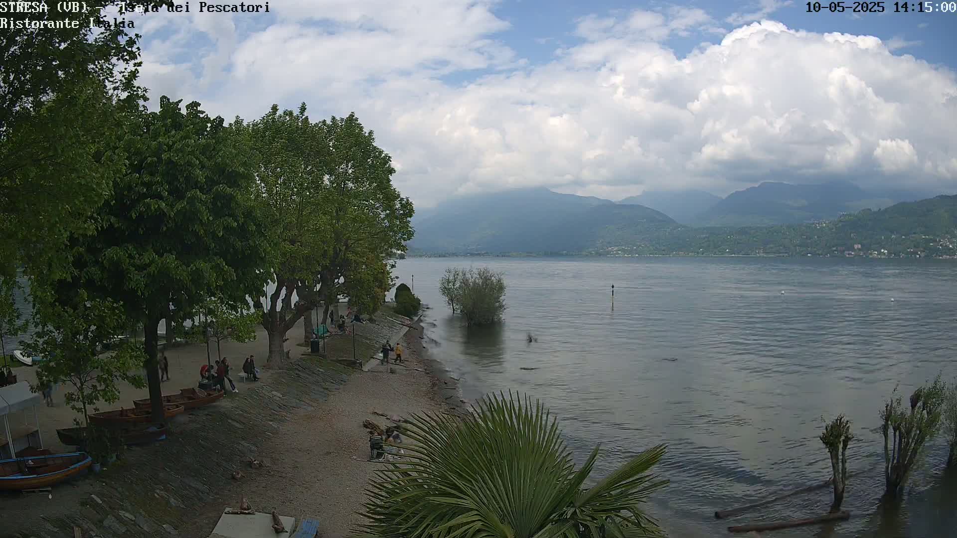 A partly cloudy day shows a lakeside view with people relaxing on a pebbled beach near some trees and small boats, mountains in the distance.