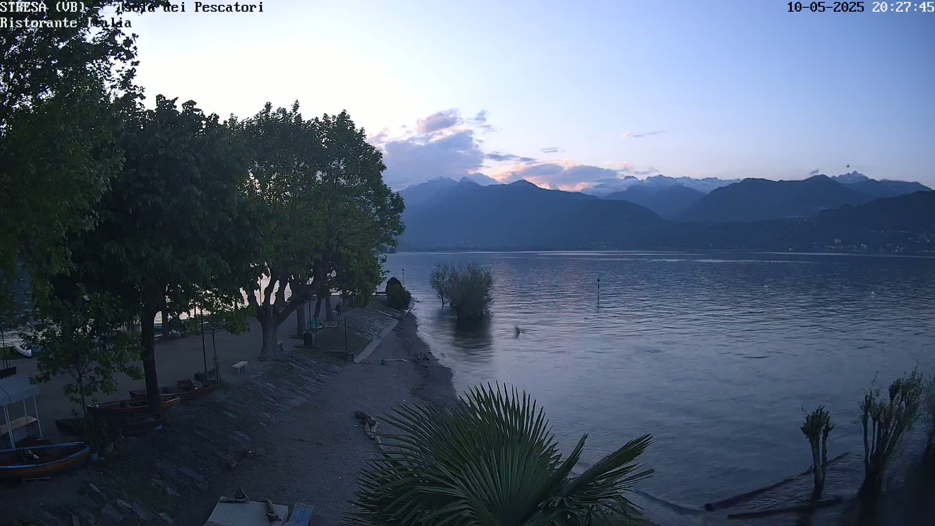 A calm lake at dusk, with a small beach, trees, and boats, is backed by mountains with some snow visible under a partly cloudy sky.