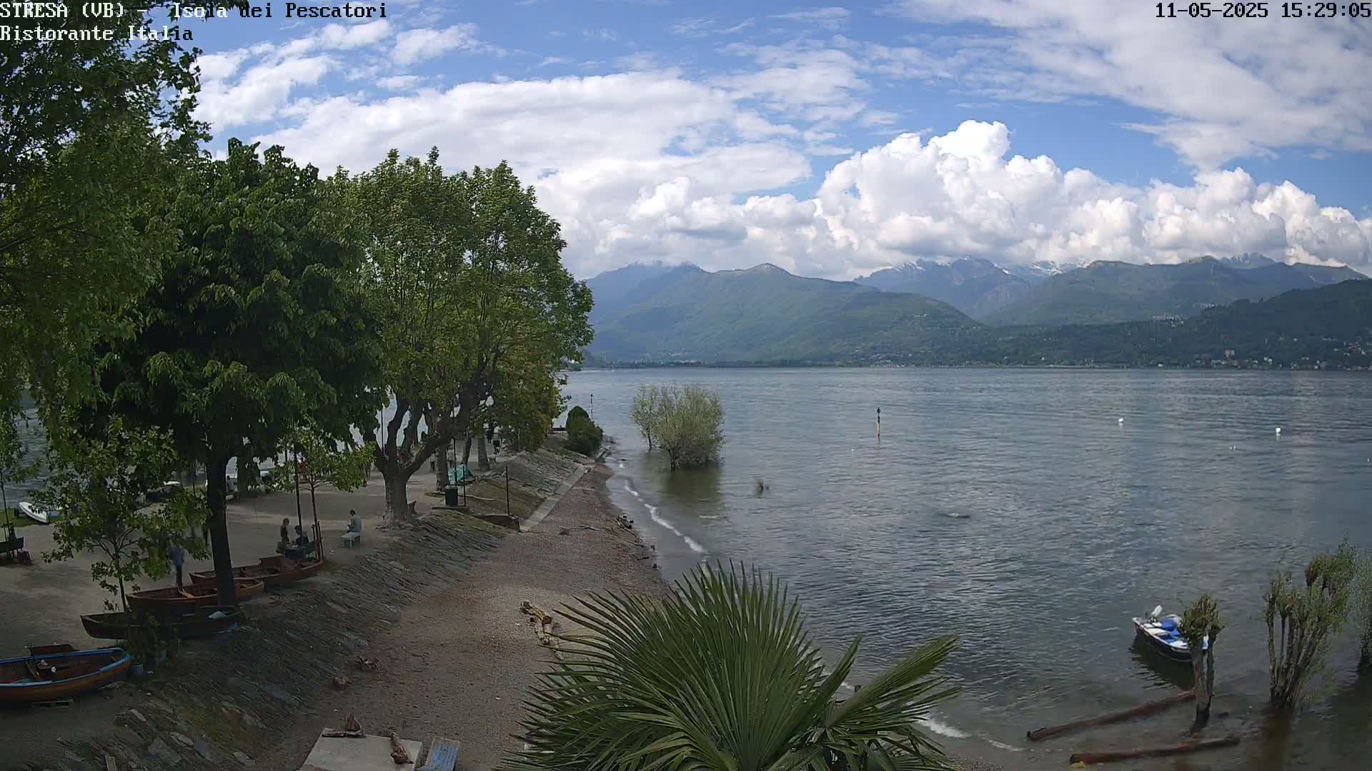 A partly cloudy day reveals a calm lake with mountains in the background, a small beach with several boats, and lush green trees lining the shoreline.