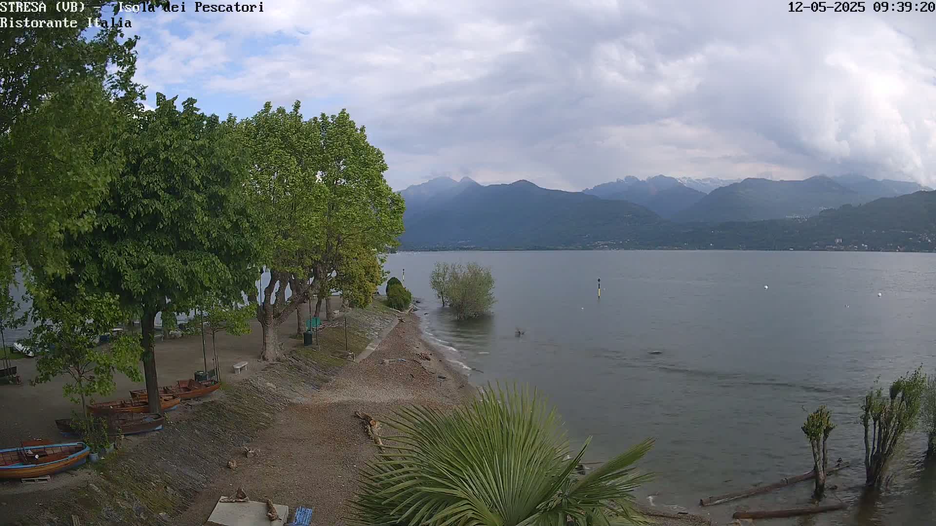 A lake shore with several small boats pulled up on a pebble beach, lined with leafy green trees, under an overcast sky, with mountains in the distance.