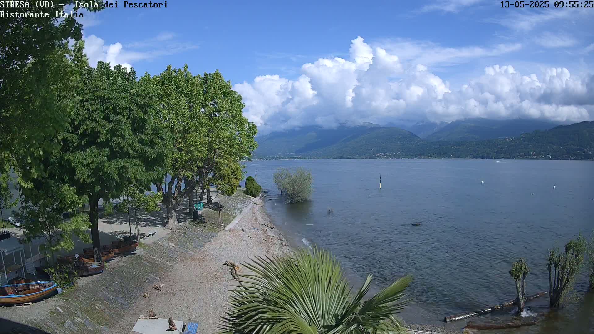 A partly cloudy sky overlooks a calm lake, mountains in the distance, a pebble beach with several small boats, and lush green trees.