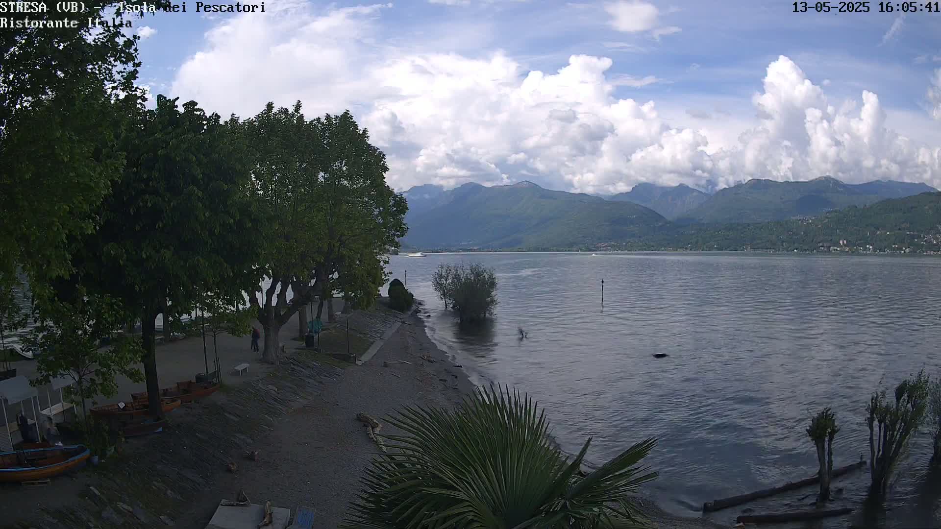 A partly cloudy day reveals a lake shore with trees, a pebble beach, small boats, and distant mountains.