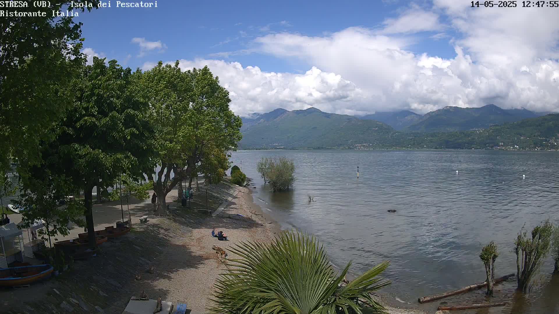 A partly cloudy day shows a view of a lake with mountains in the background, a sandy beach with a few people, and lush green trees along the shoreline.