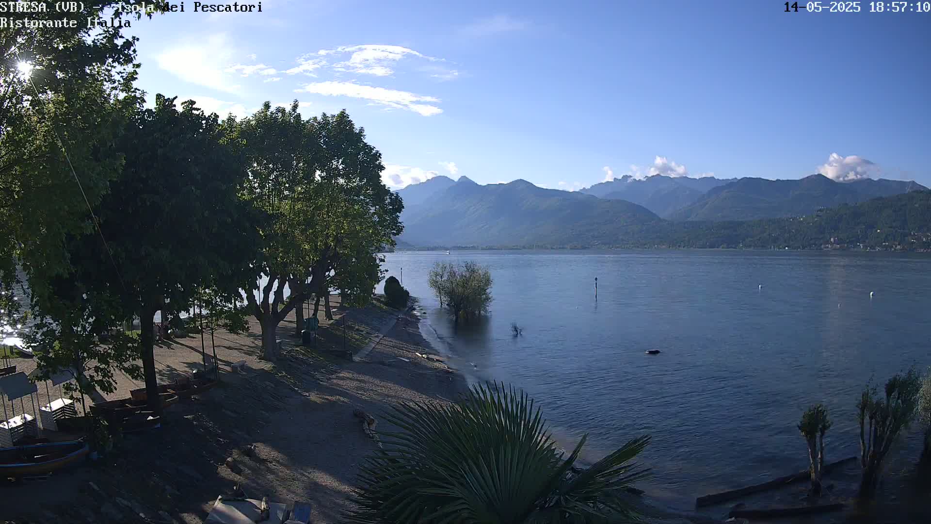 A calm lake with mountains in the background is viewed from a shoreline with trees, a small beach, and some boats.