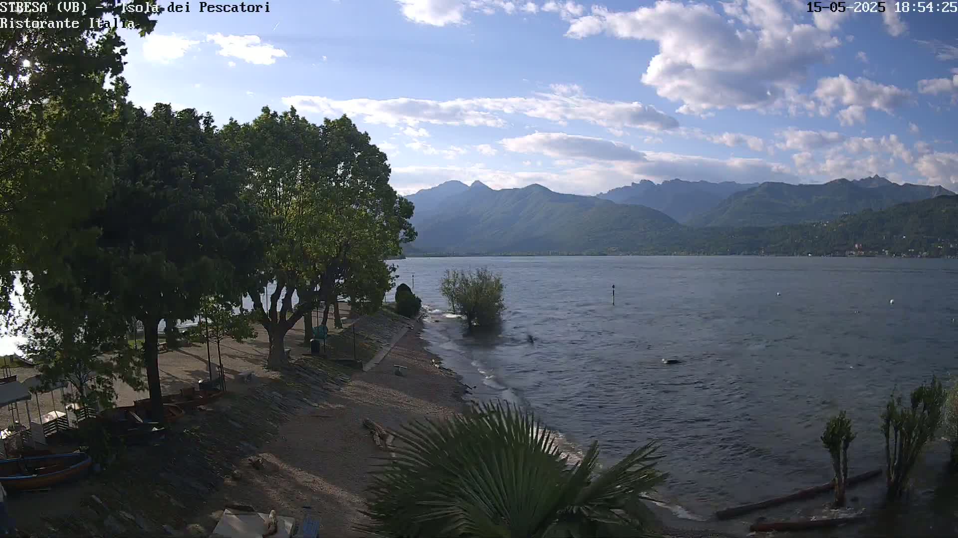 A partly cloudy day shows a lake with small waves lapping a sandy shore lined with trees, under a partly cloudy sky with mountains in the background.