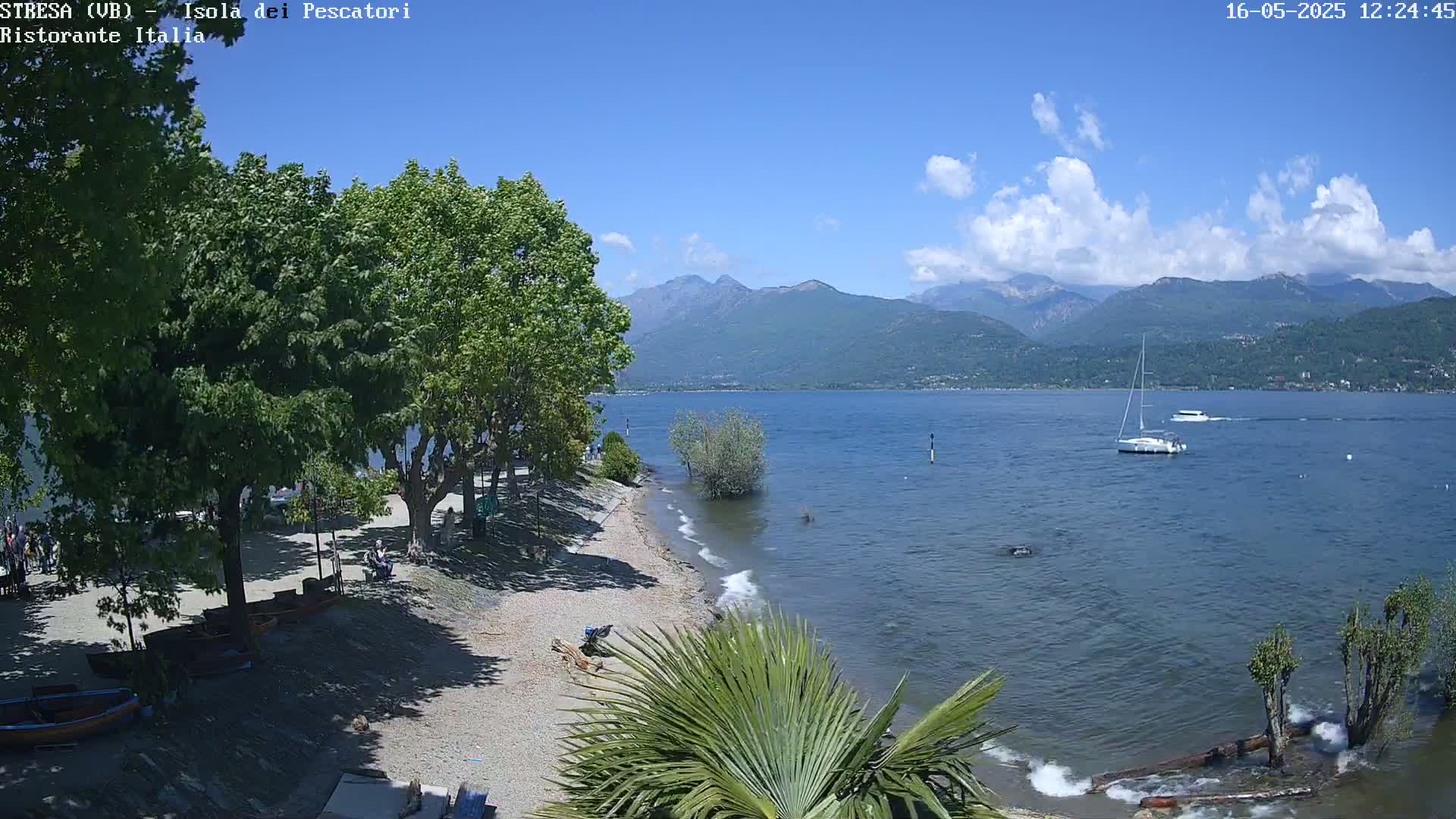 A sandy beach lined with trees and boats sits on the edge of a calm lake under a mostly sunny sky, with mountains visible in the distance.