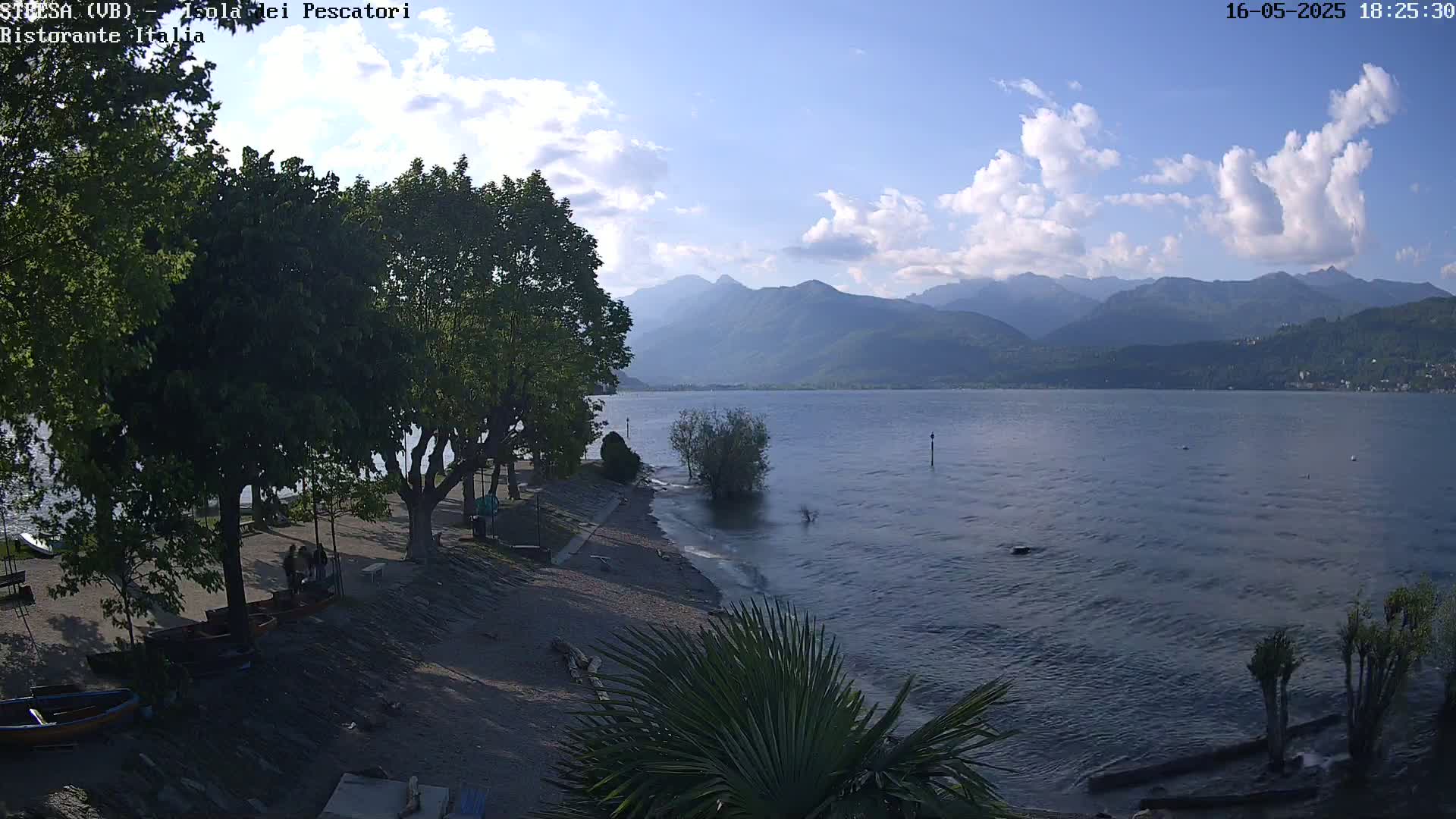 A partly cloudy day overlooks a calm lake with mountains in the background, a sandy shoreline with trees and several small boats.