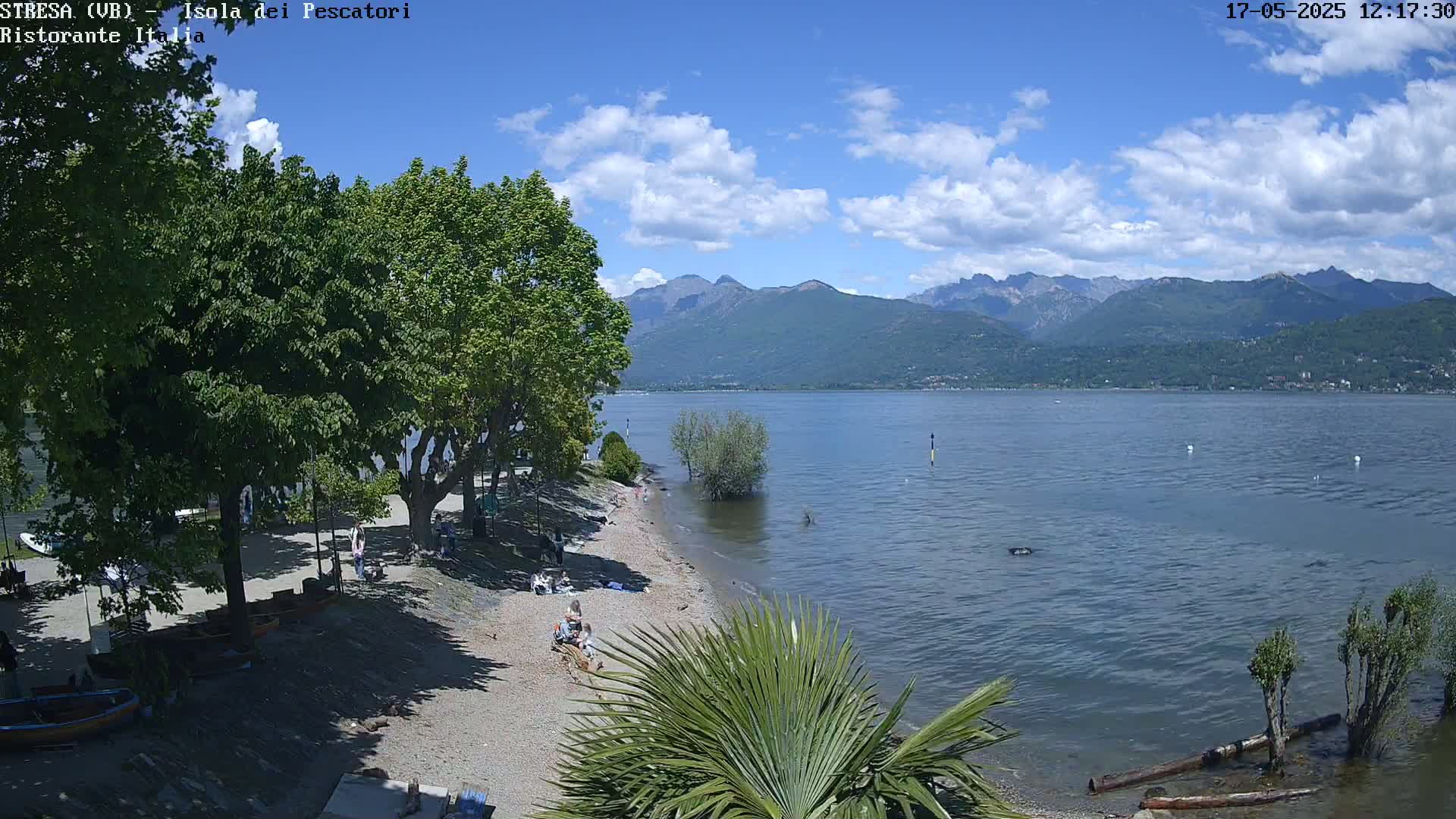 A partly sunny day shows a pebble beach with people relaxing near a calm lake, backed by lush green trees and distant mountains.