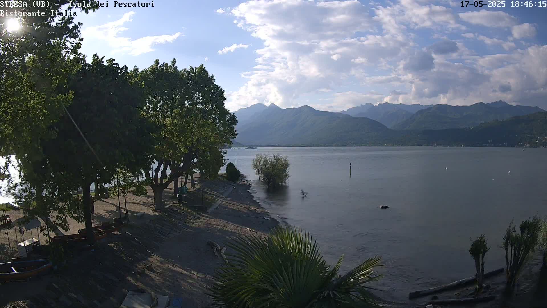 A partly cloudy day shows a lake with mountains in the background, a sandy shoreline with trees and some boats, and a few palm trees in the foreground.