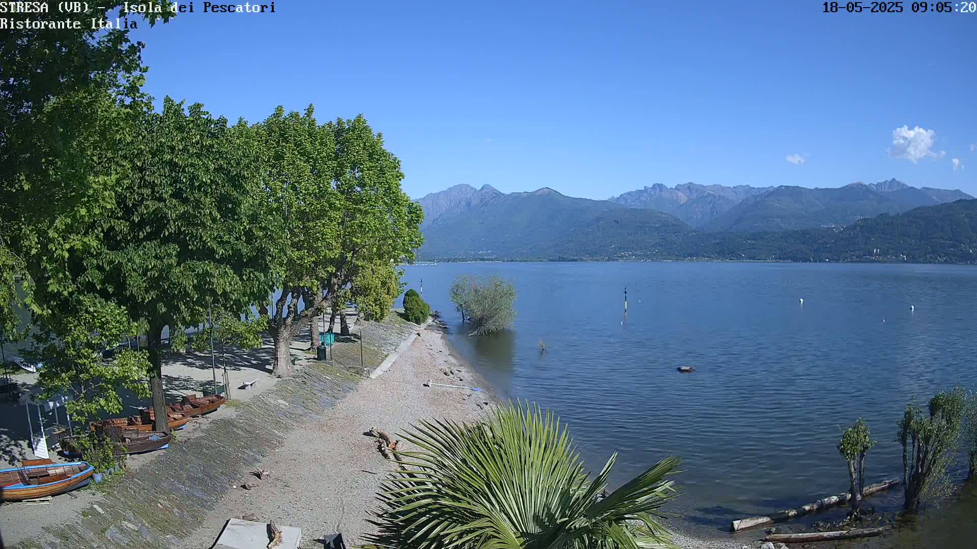 A calm lake with mountains in the background, under a clear blue sky, is seen from a lakeside beach with trees and several small boats.
