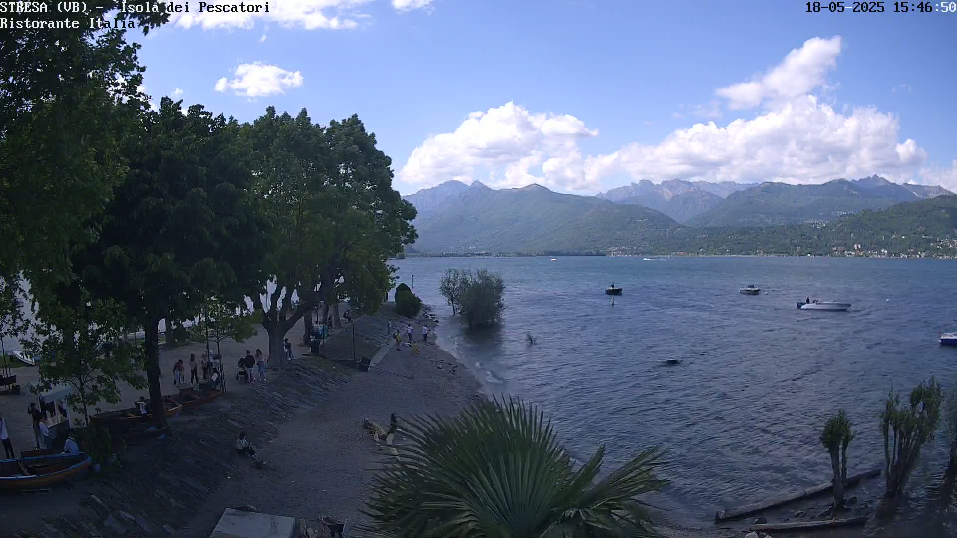 A partly cloudy day shows a lakeside view with people gathered on a pebbled beach near trees, several small boats on the water, and mountains in the distance.