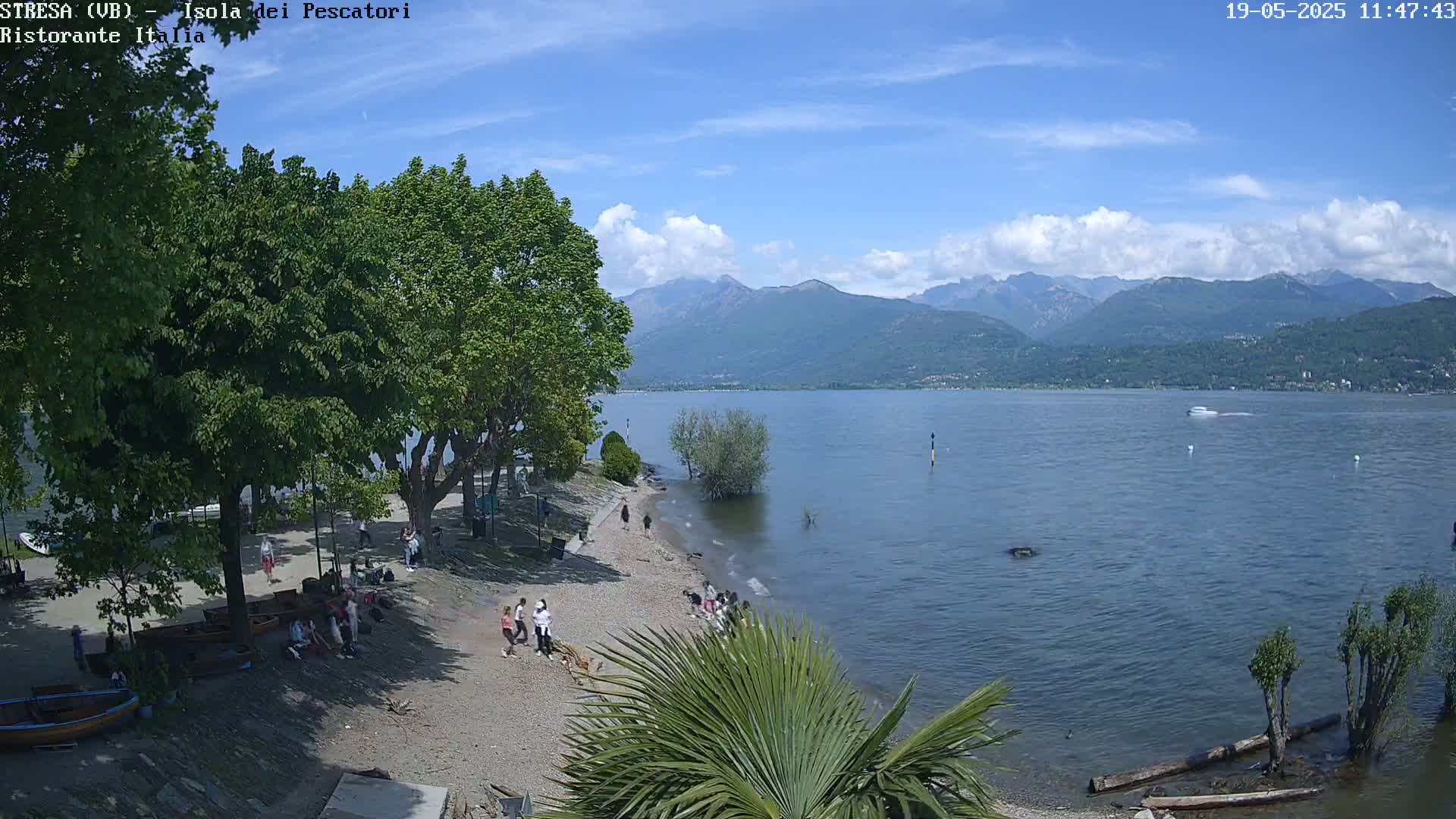 A sunny day on a pebble beach, with people relaxing and walking near the water's edge, overlooking a calm lake and mountains in the distance.