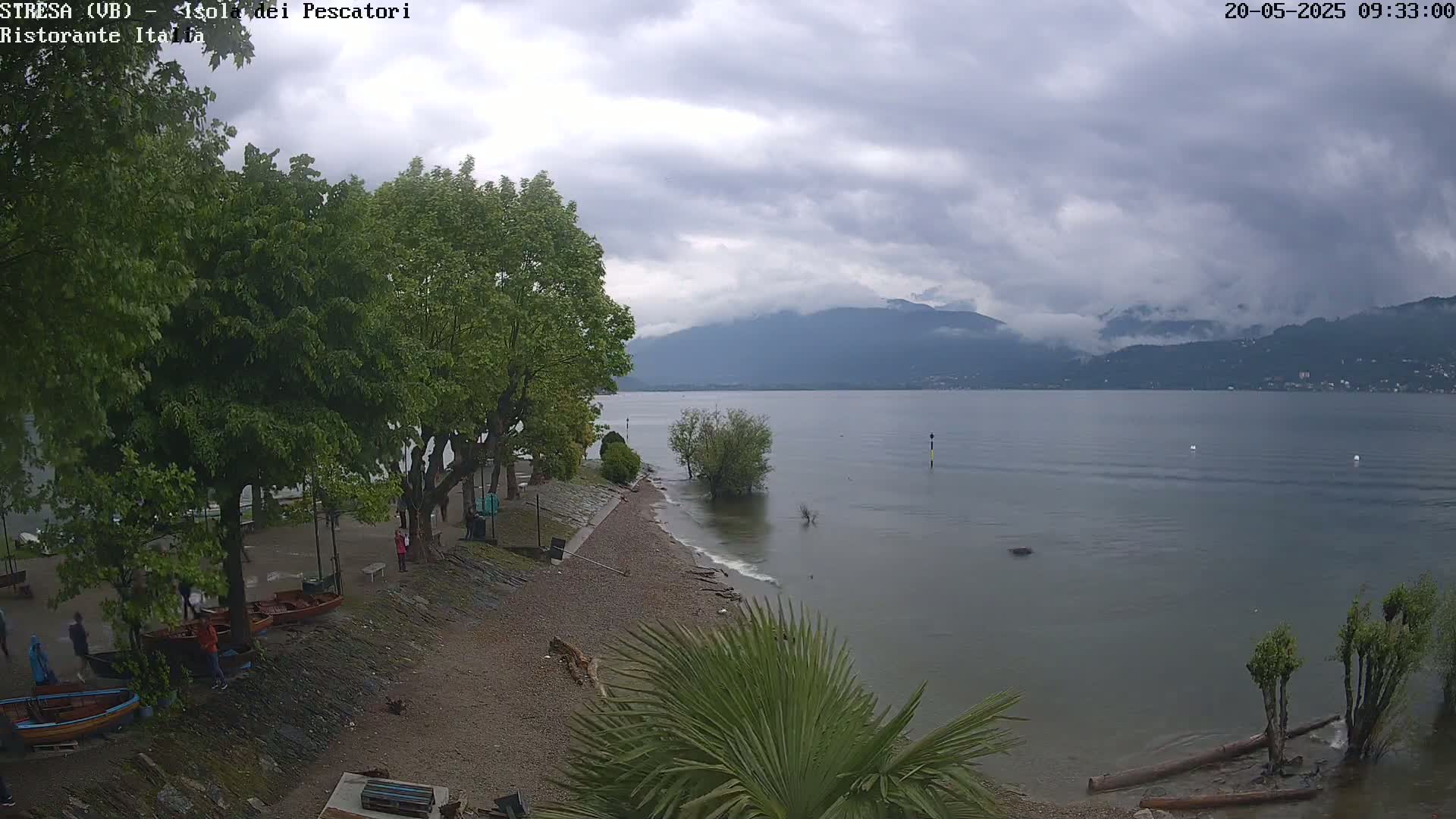 A partly cloudy day overlooking a calm lake with mountains in the distance, a gravel shoreline with several people and small boats, and lush green trees.