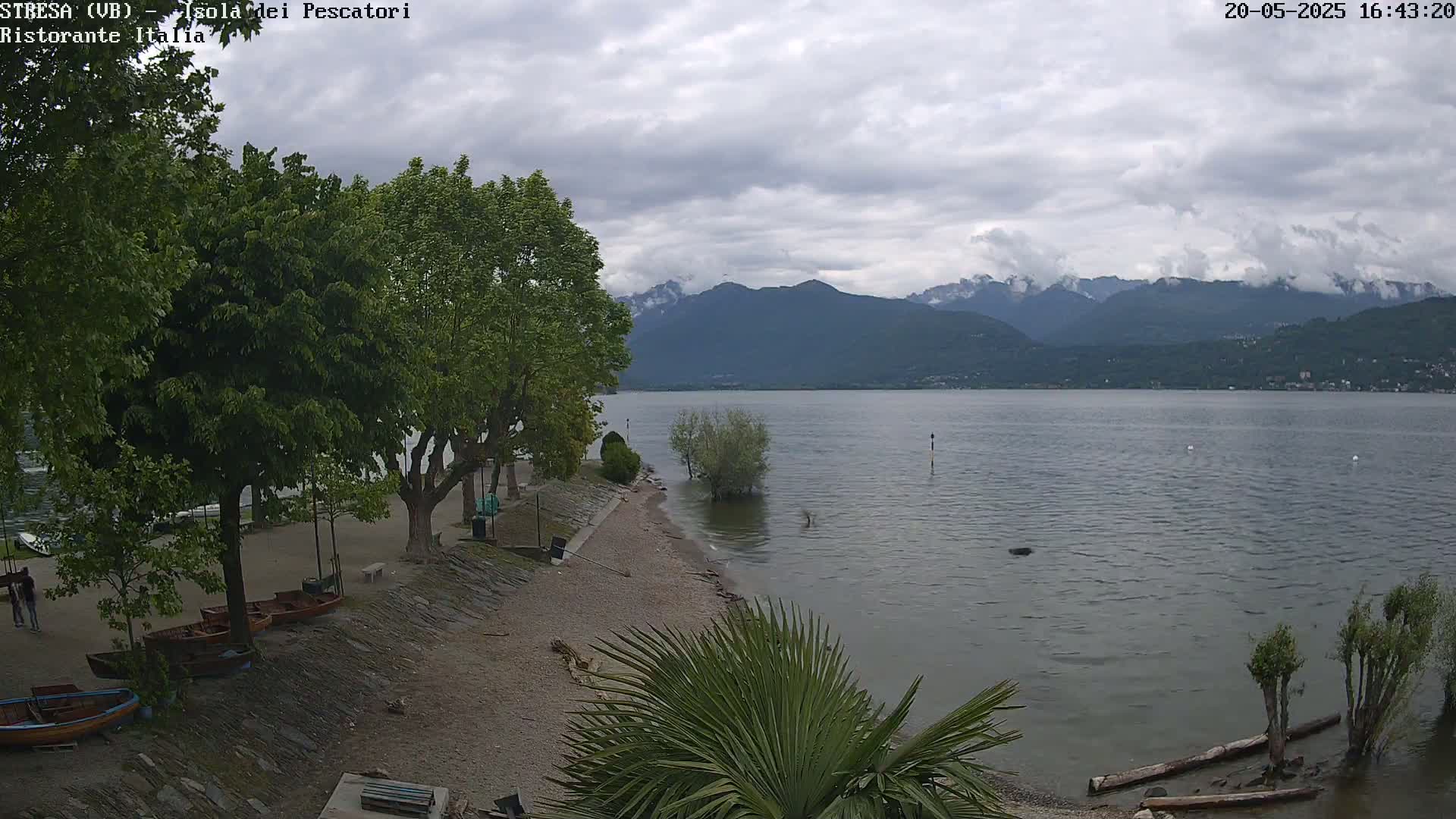 A calm lake stretches out before a pebbled shoreline lined with trees and several small boats, under an overcast sky and mountains in the distance.