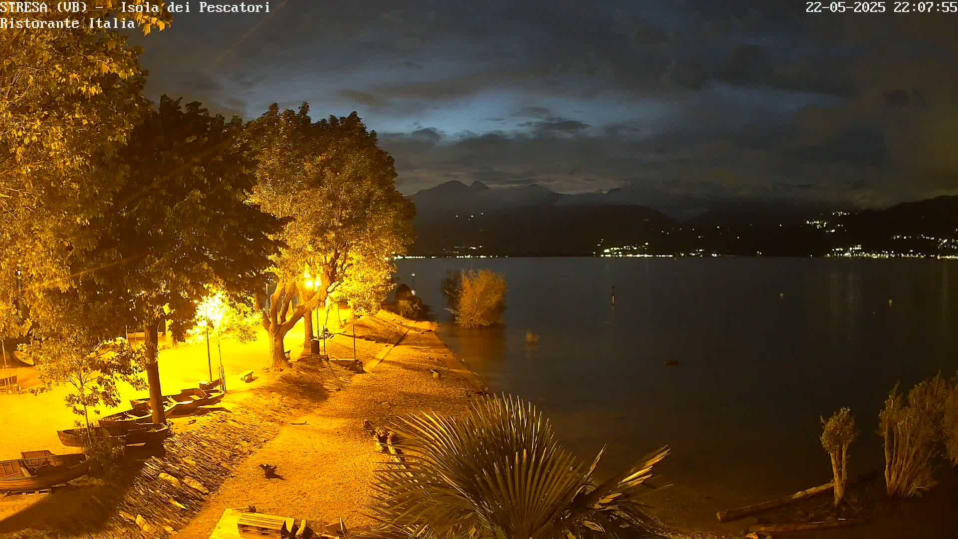 A nighttime view of a calm lake under a partly cloudy sky, with illuminated trees, a shoreline path, and several small boats docked on a sandy beach.