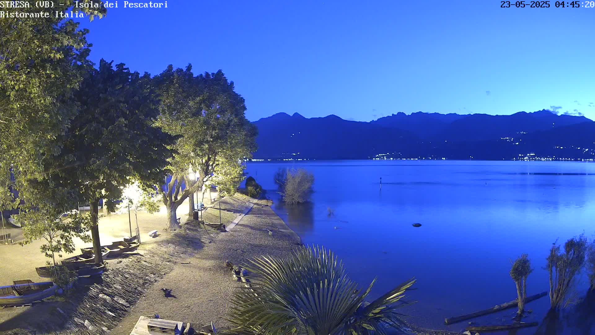 A tranquil nighttime scene shows a calm lake reflecting the dark blue twilight sky, mountains in the distance, and several boats resting on a shoreline lined with trees and shrubs.