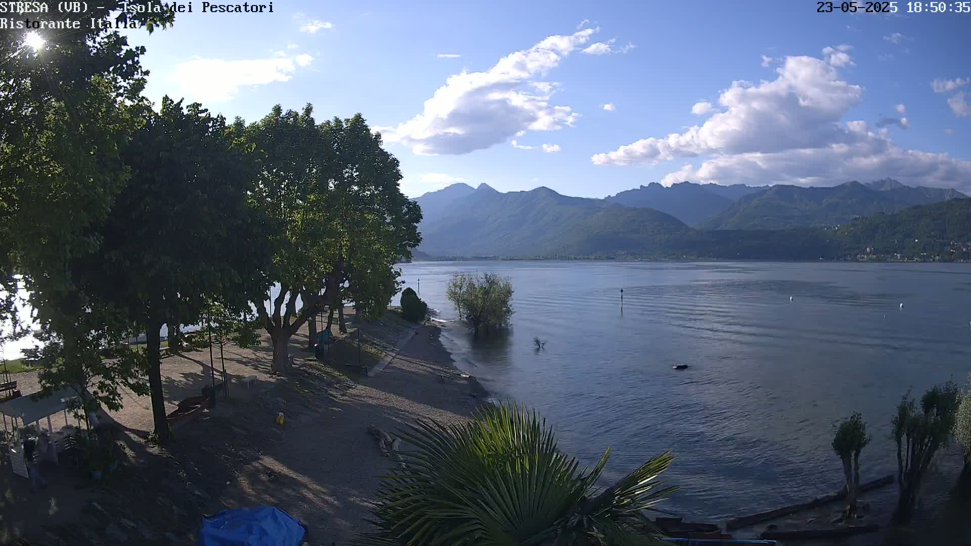 A partly sunny day shows a sandy lakeshore with trees, leading to a calm lake and distant mountains.