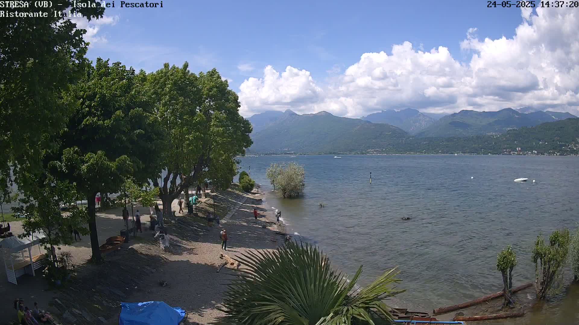 A partly cloudy day reveals a lake shore with people relaxing on a sandy beach near lush green trees and mountains in the distance.