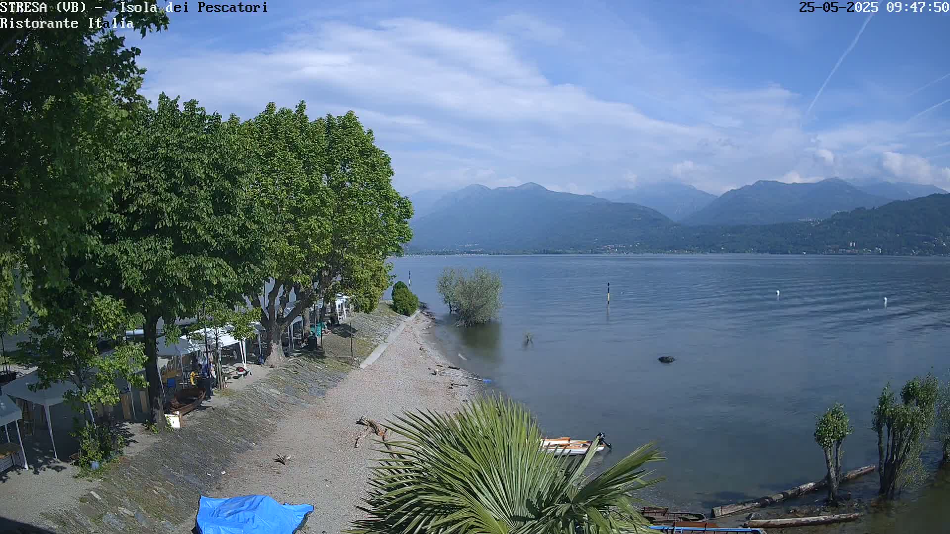 A partly sunny day shows a calm lake with mountains in the background, a sandy beach with trees and small boats, and a few people near some shaded areas.