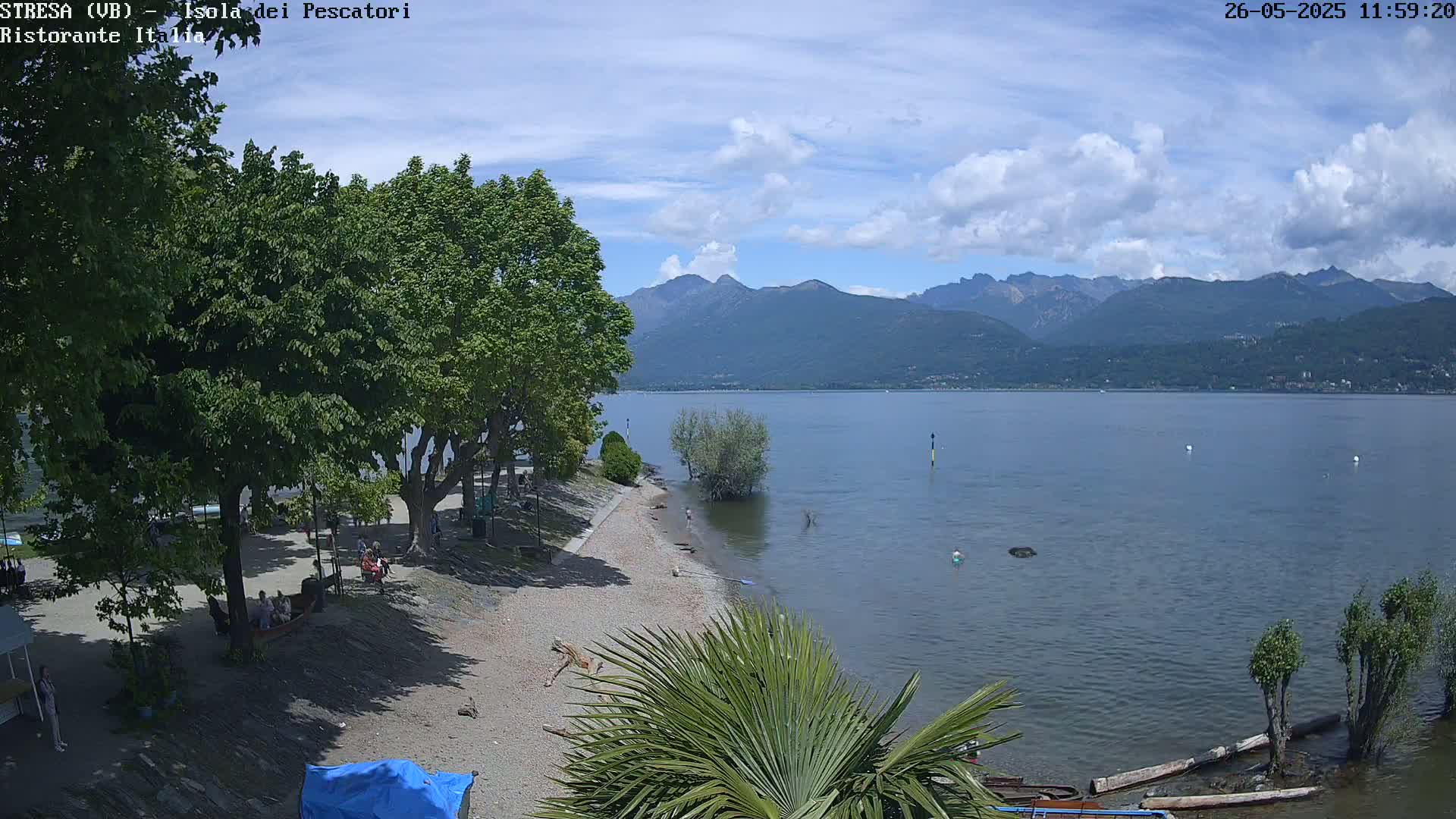 A partly sunny day shows a calm lake with mountains in the background, a sandy beach with people and trees lining the shore.
