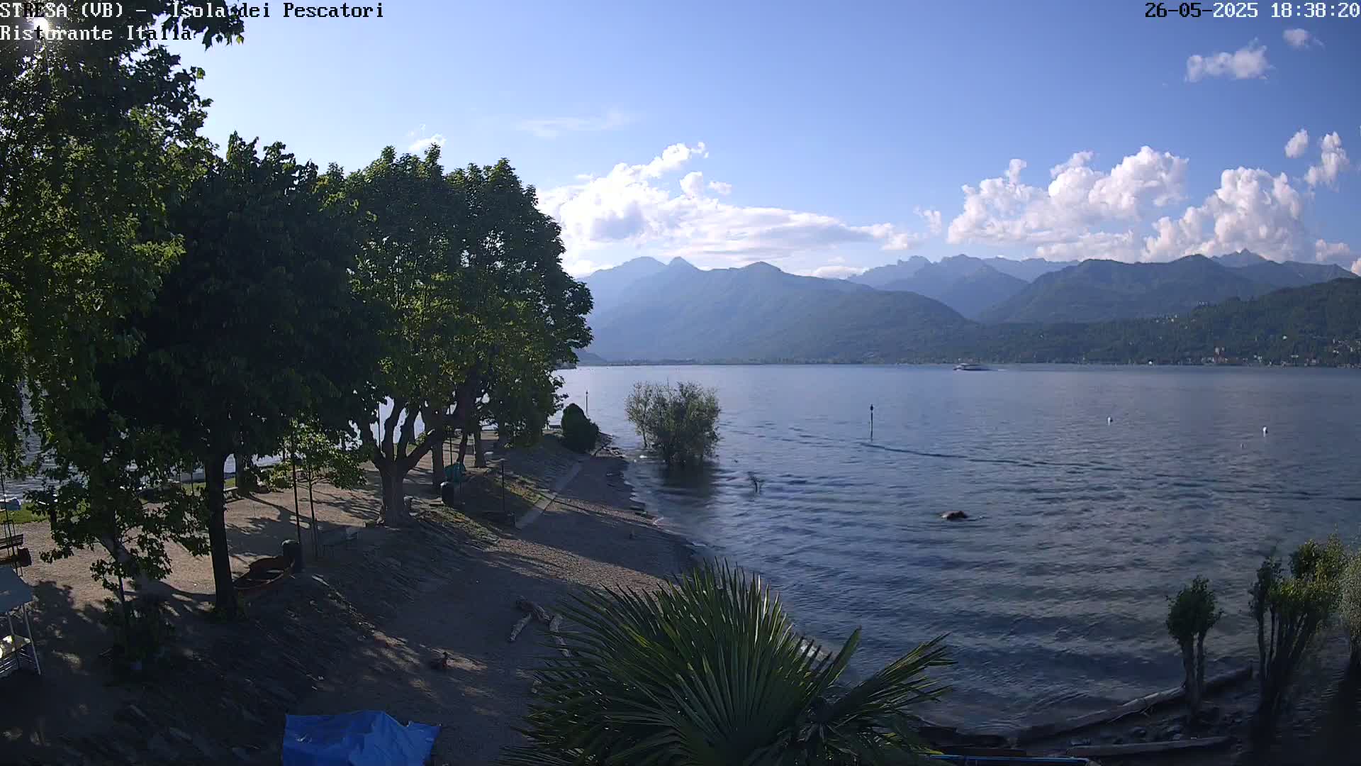 A partly cloudy day shows a calm lake with mountains in the background, viewed from a lakeside area with trees, a sandy shore, and some small boats.