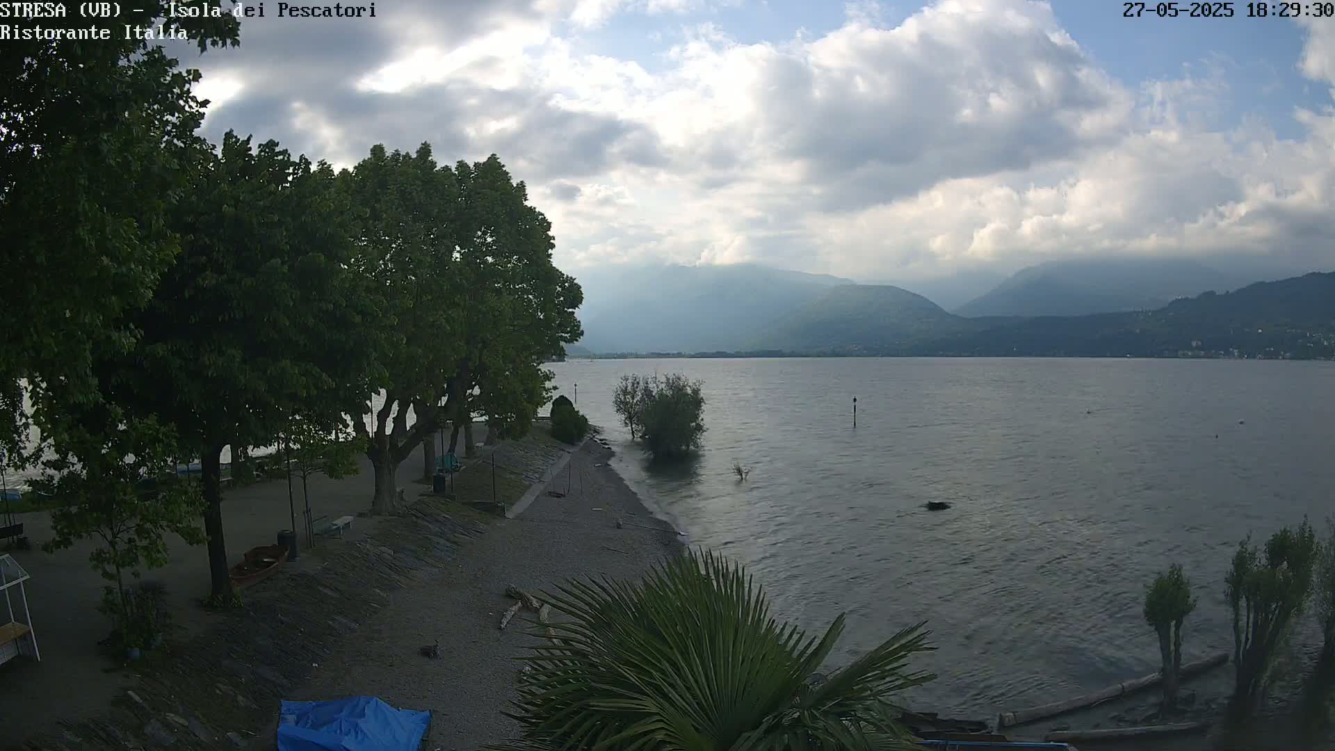 A calm lake with mountains in the background, under a partly cloudy sky, is viewed from a lakeside area with trees and a small beach.