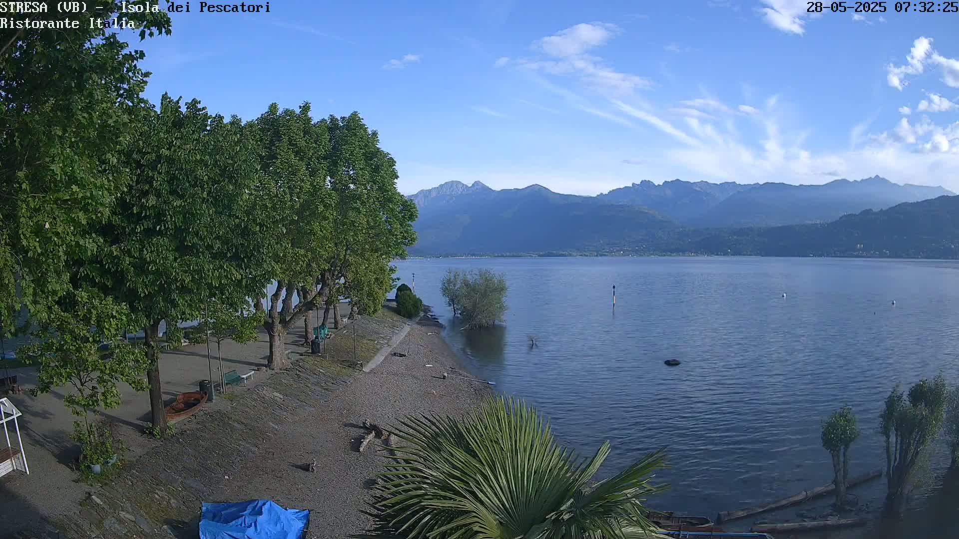 A tranquil lake shore scene under a mostly clear sky shows a pebbled beach lined with trees, a few boats, and mountains in the distance.