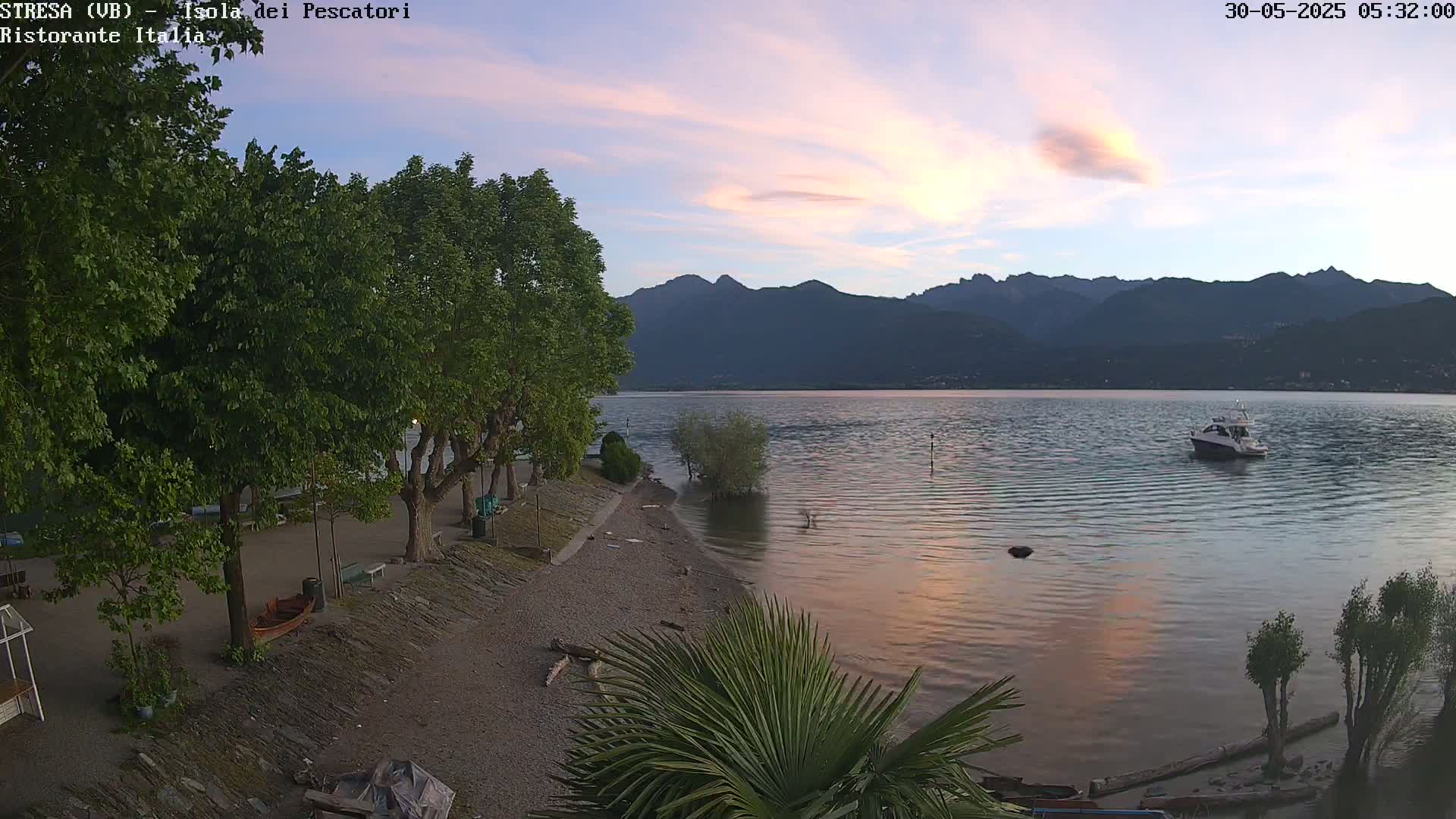 A calm lake at dawn, with a motorboat on the water,  is bordered by a pebbled shoreline with trees and a small, rocky beach, and mountains are visible in the background under a pastel-colored sky.