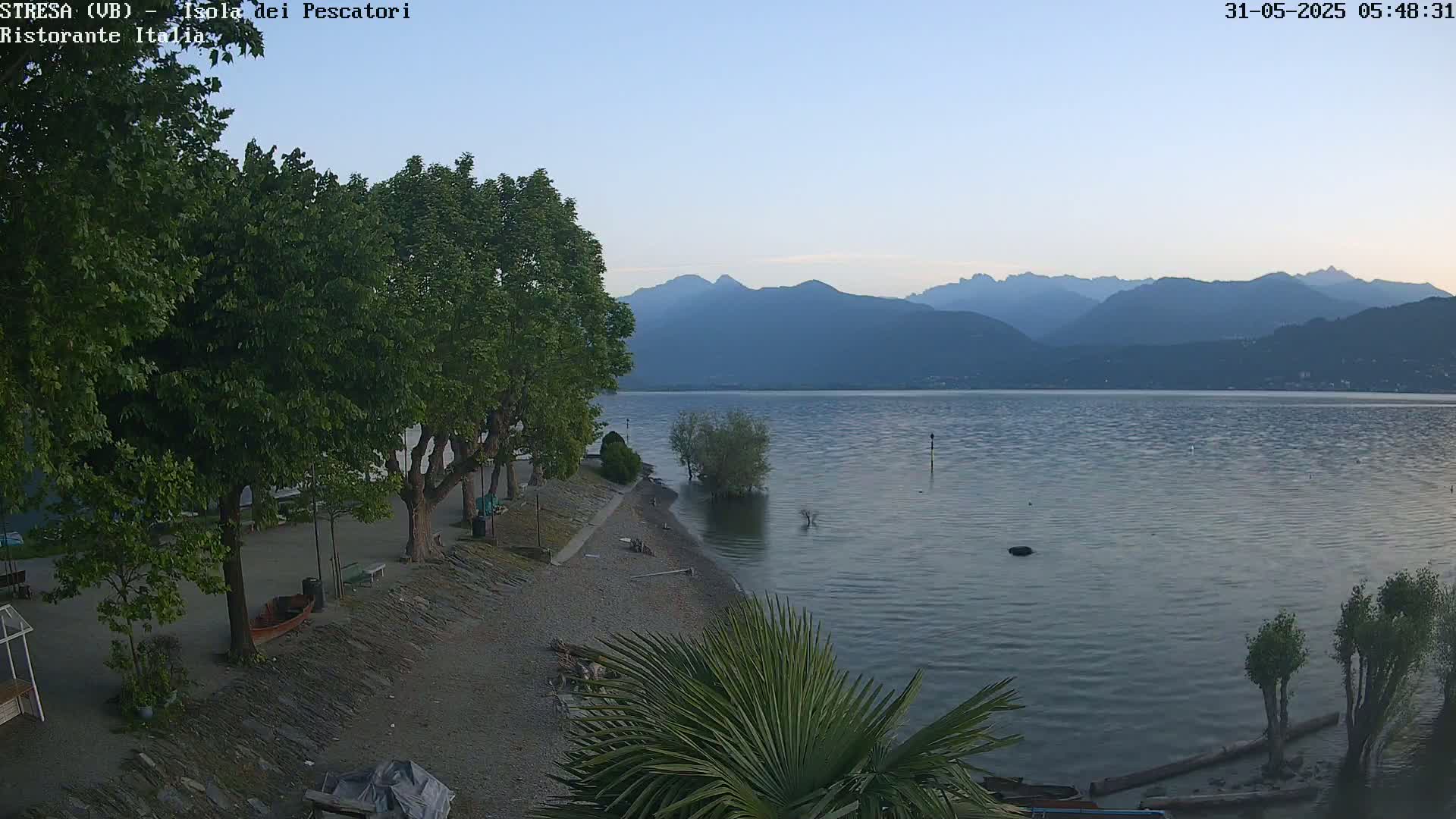 A calm lake is seen at dusk, with mountains in the background and a tree-lined shoreline featuring a small sandy beach.