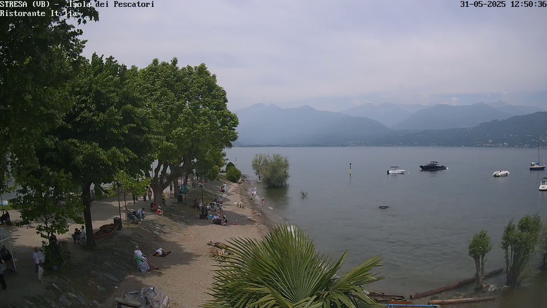 A hazy, overcast day reveals a lake shore with people relaxing on a sandy beach beneath leafy trees, several boats on the calm water, and mountains in the background.