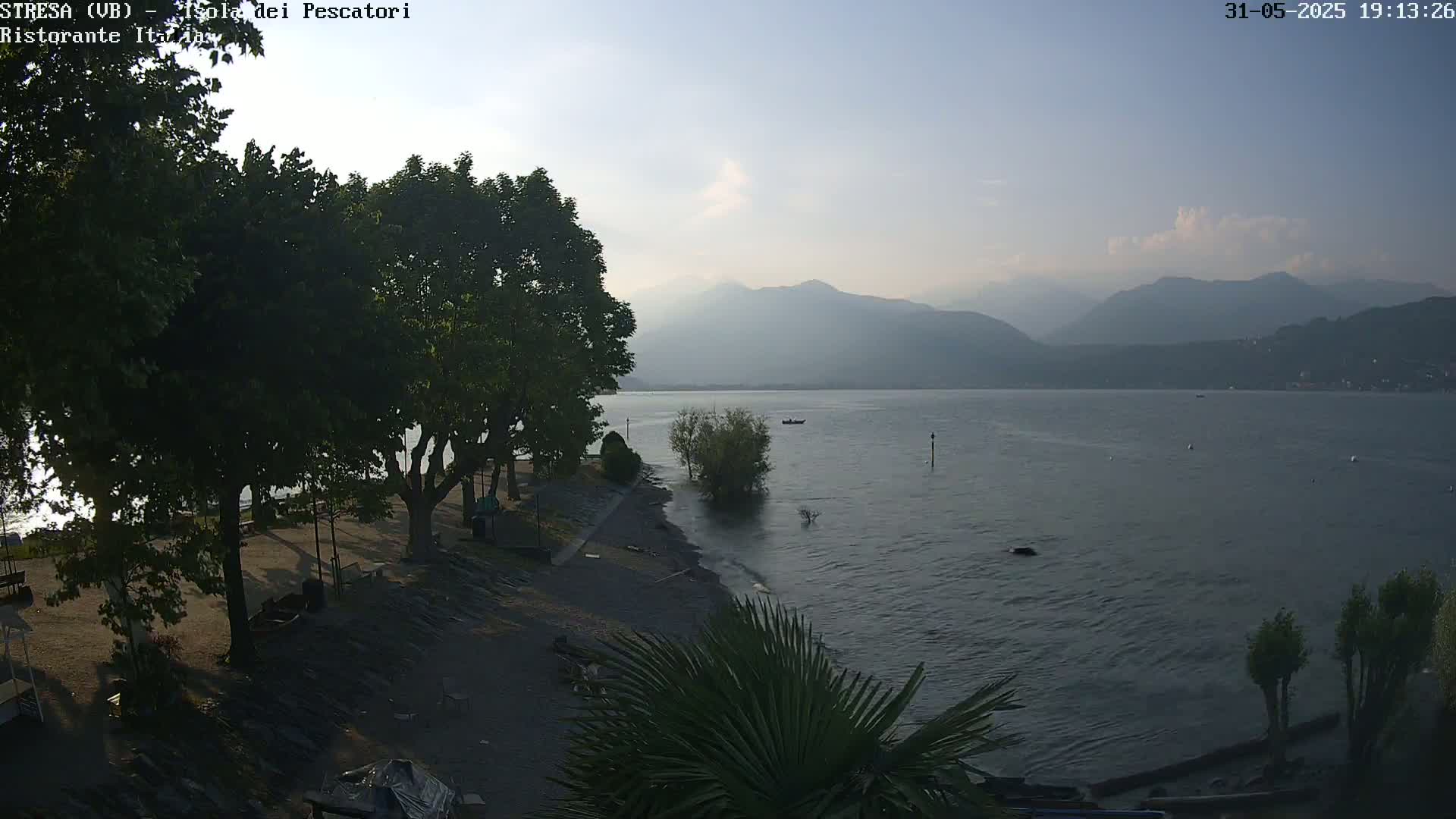 A calm lake with hazy mountains in the background is seen from a lakeside park with leafy trees, under a mostly clear sky.