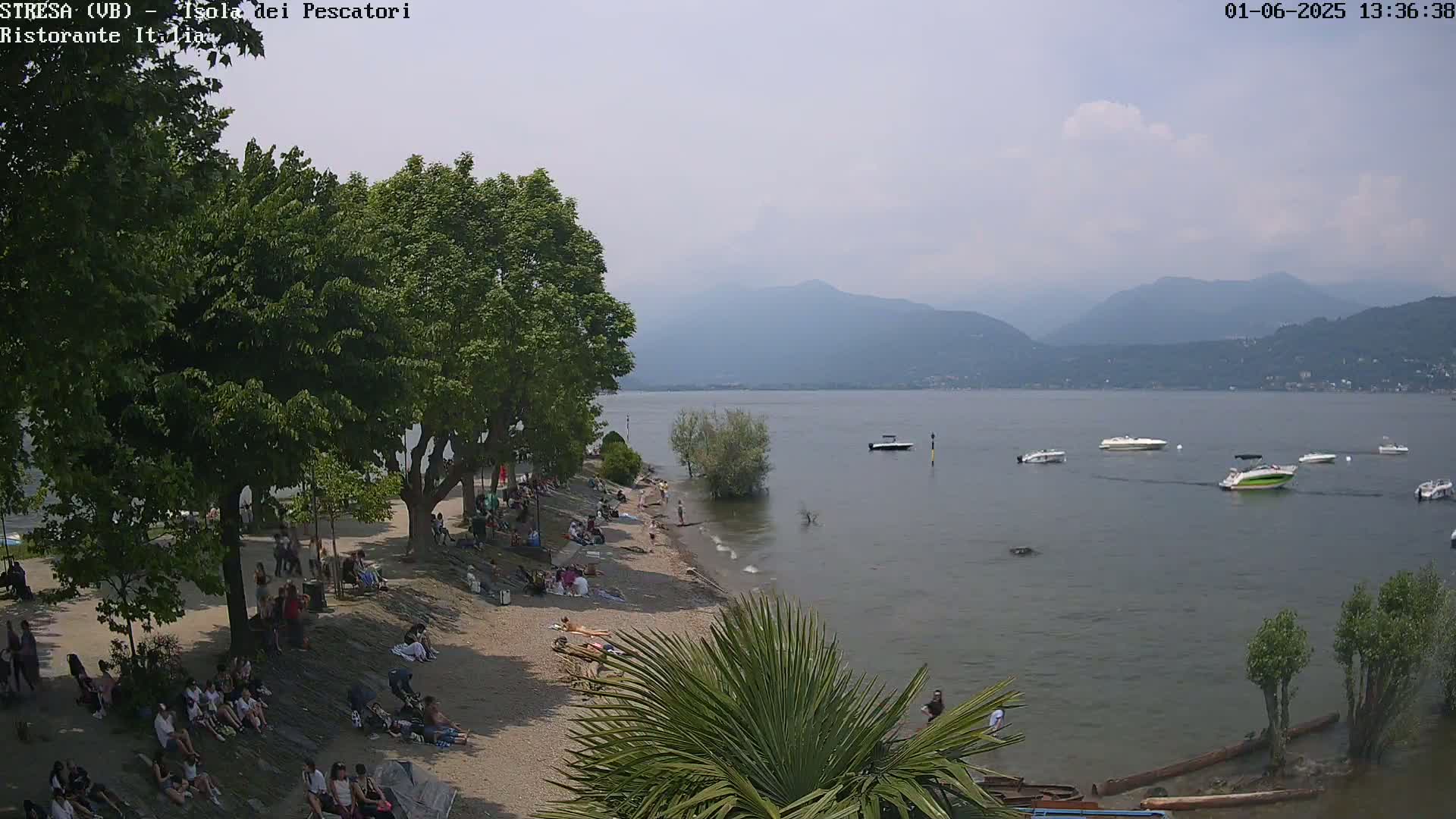 A sunny day at a lakeside beach shows people relaxing under shady trees and on a sandy shore, with several motorboats on the calm water and mountains in the background.