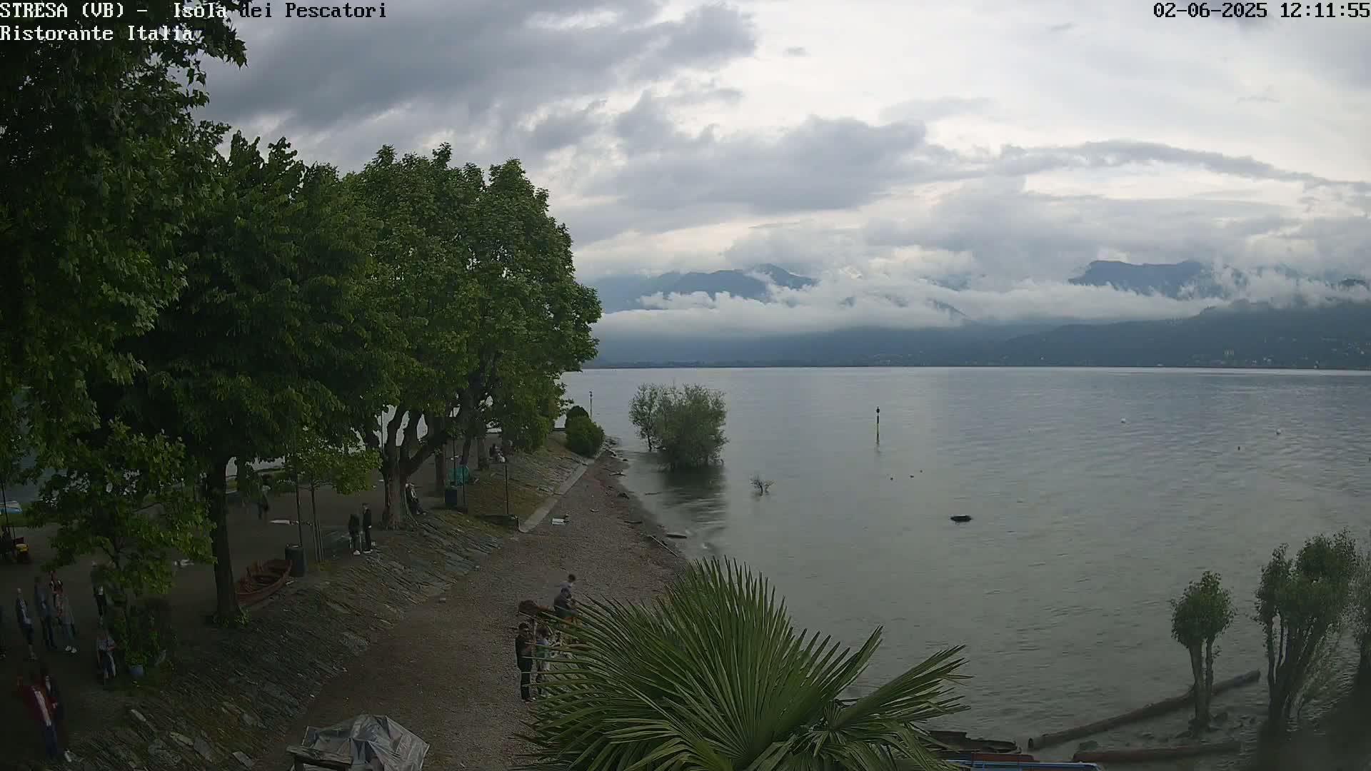 A calm lake under a cloudy sky is bordered by a stone shoreline with trees and people gathered near the water's edge.