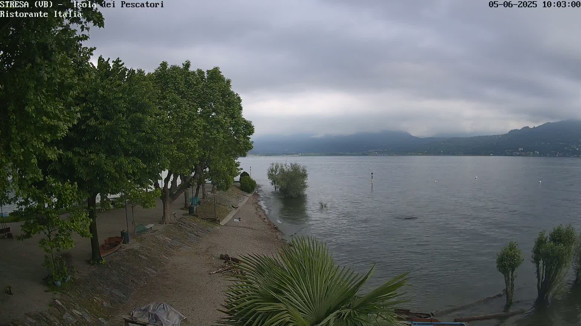 A calm lake with overcast skies is viewed from a shoreline with green trees and a sandy beach area.
