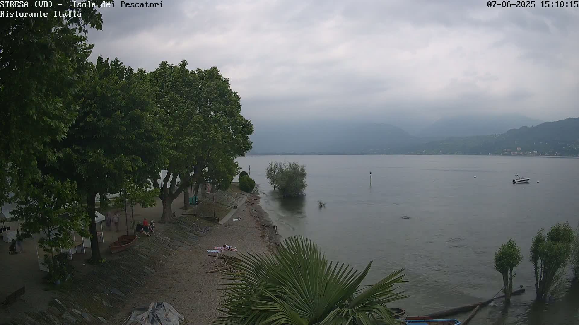 A calm lake under an overcast sky is seen from a lakeside perspective, with trees lining a sandy shore where a few people are relaxing.