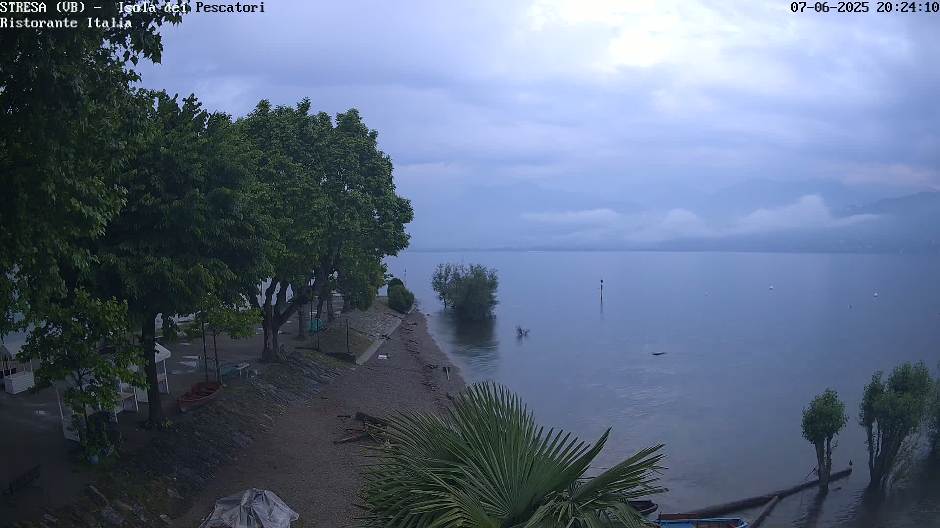 A calm lake under an overcast sky is seen from a high vantage point, with a pebble beach lined with trees and small structures in the foreground.