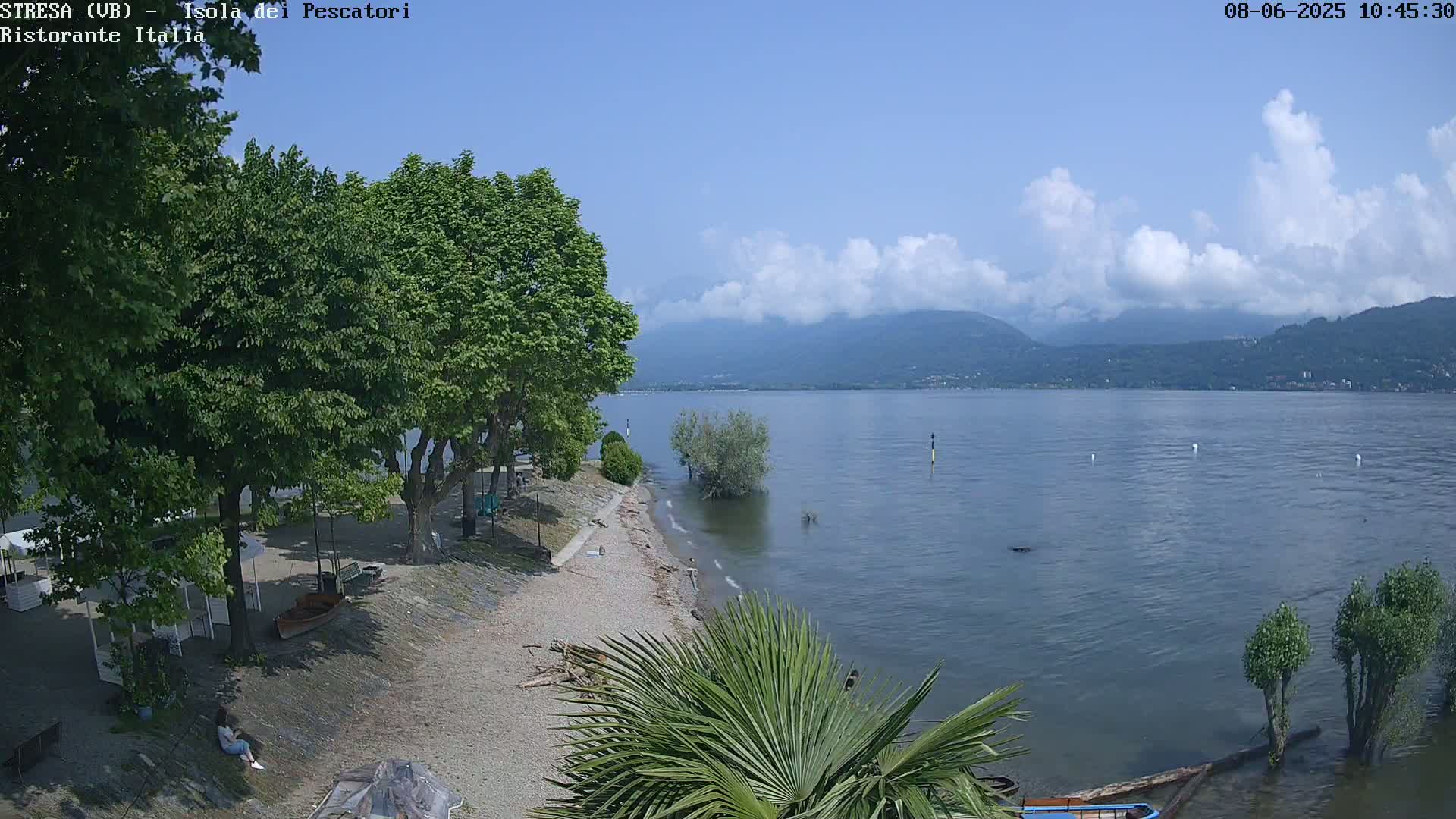 A partly sunny day shows a calm lake with mountains in the background, a sandy shoreline with trees and a small beach, and a person sitting near some small structures.