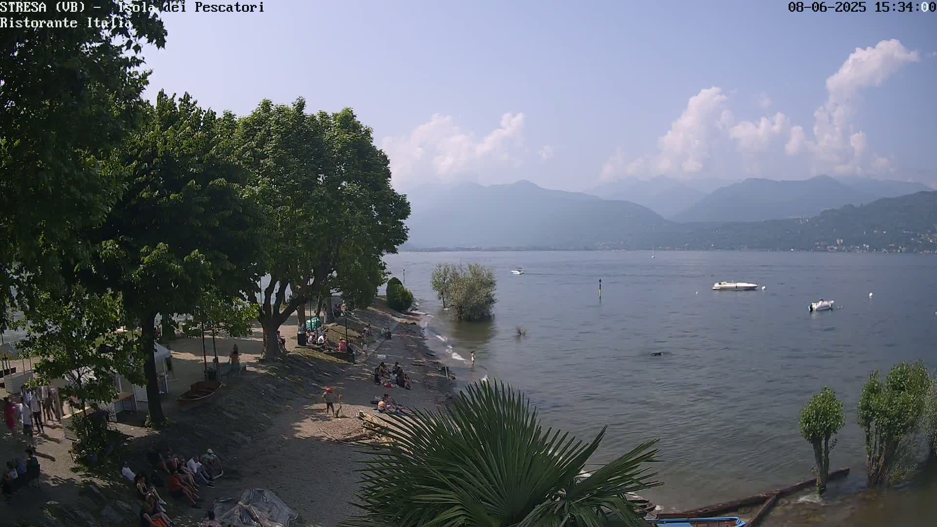 A sandy lakeshore with people relaxing and swimming, shaded by lush trees, overlooks calm lake water with distant mountains under a partly cloudy sky.