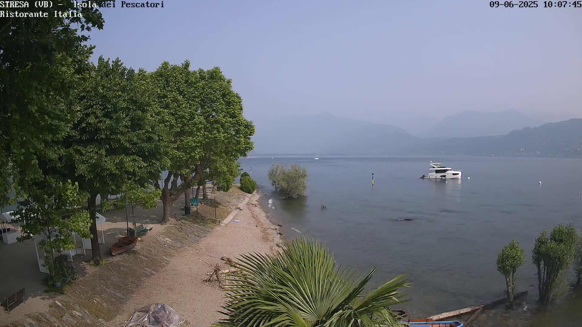 A hazy, sunny day overlooking a calm lake with a motorboat, a sandy shoreline lined with trees, and a small beach area.