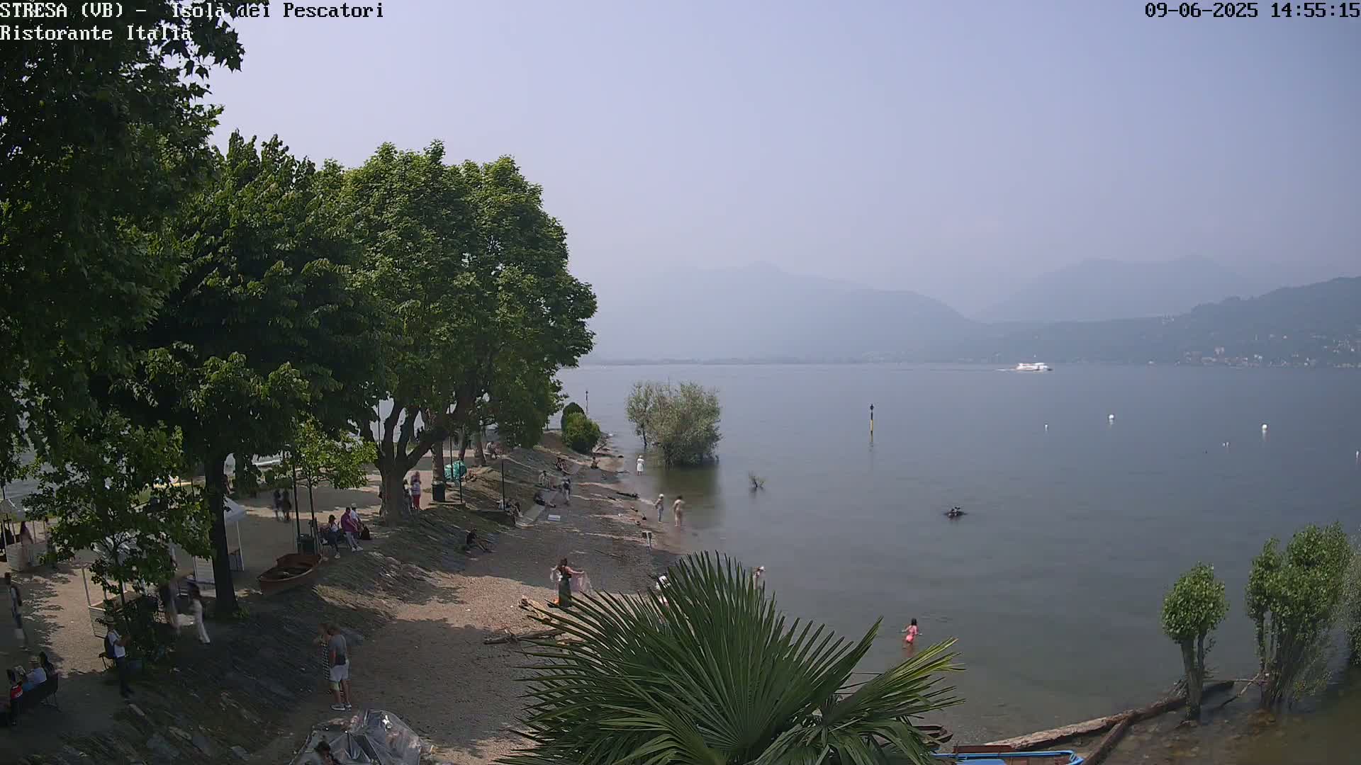 A hazy view of a lake shore with people relaxing on a pebble beach, shaded by trees, and mountains visible in the distance.
